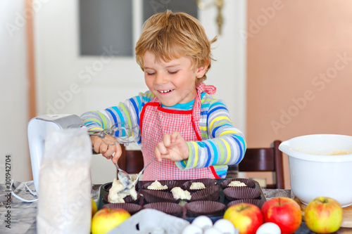 Cute little happy blond preschool kid boy baking apple cake and muffins in domestic kitchen. Funny lovely healthy child having fun with working with mixer, flour, eggs, fruits. Little helper indoors
