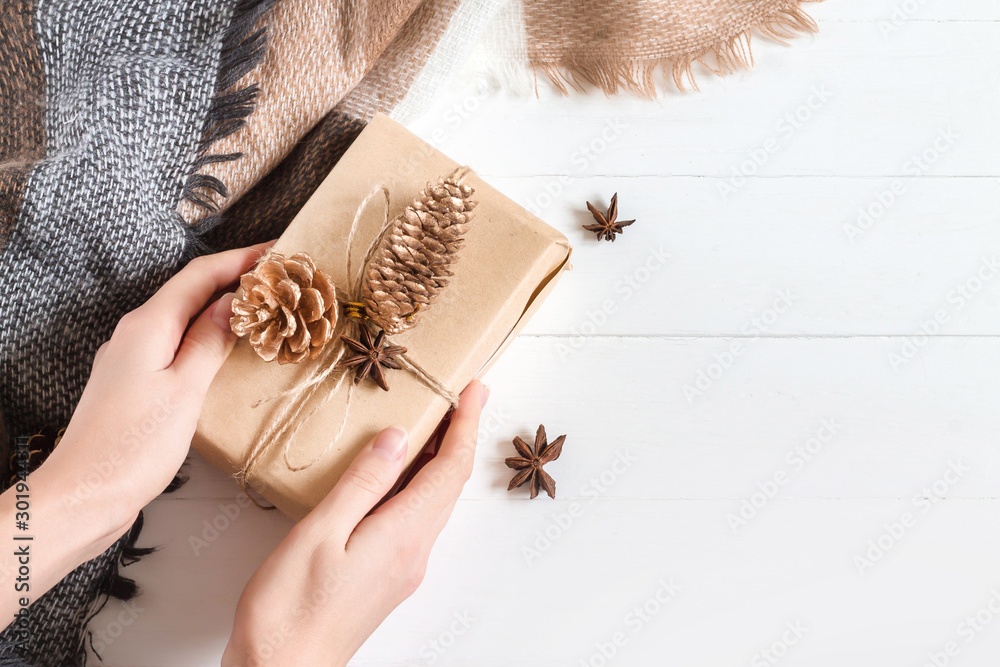 Obraz premium Female hands hold a gift box in craft paper with anise, dry cones, orange and cinnamon stick on a white wooden background in rustic style, copy space, top view