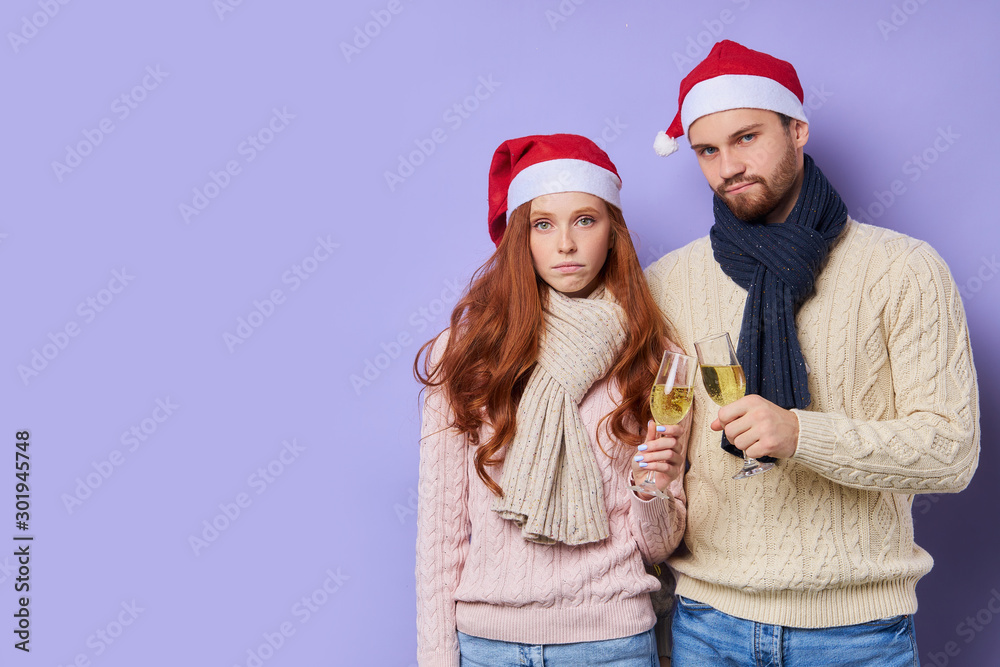 Sad upset looking couple standing with glass of champagne in hands, wearing Santas hat, looking sad, having boring party, tired from celebration, family xmas, isolated on violet background Sad upset looking couple standing with glass of champagne in hands, wearing Santas hat, looking sad, having boring party, tired from celebration, family xmas, isolated on violet background