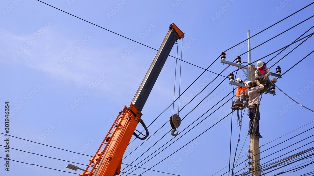 Low angle view of electricians team with crane are working to install electrical system on electric power pole against blue sky background, technology and development concept Low angle view of electricians team with crane are working to install electrical system on electric power pole against blue sky background, technology and development concept