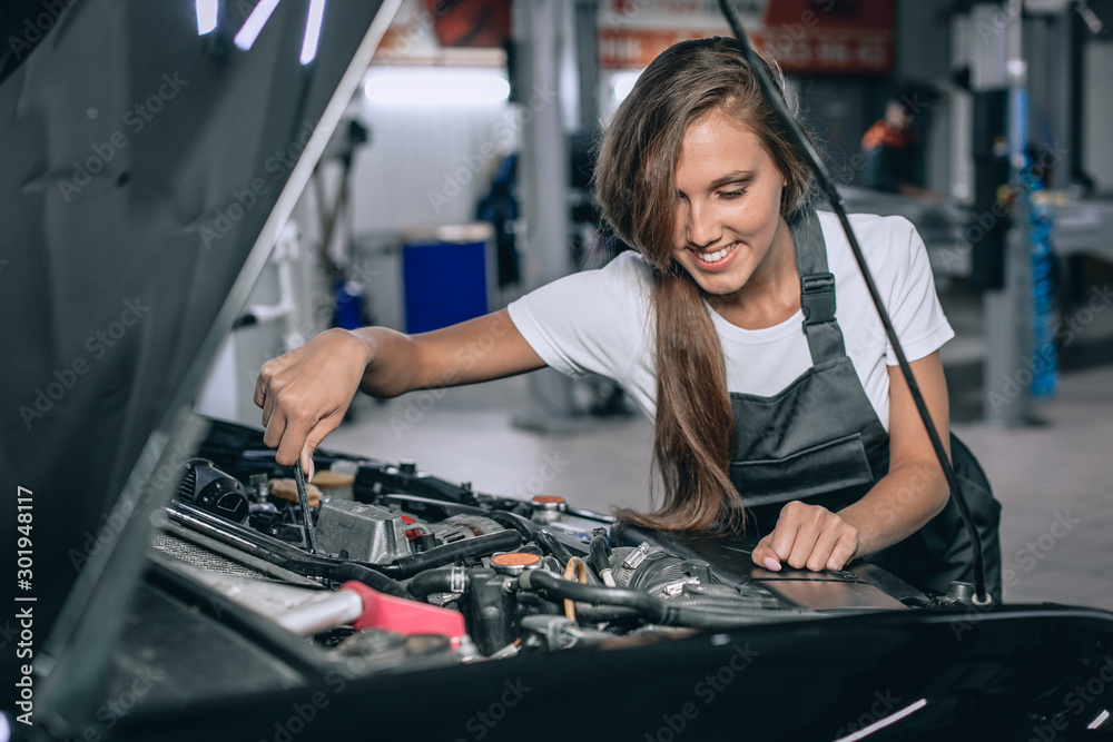 A brunette in a black jumpsuit and a white t-shirt near the open hood of black car. Young female in the garage is smiling at the camera and lowered gaze. car repair concept