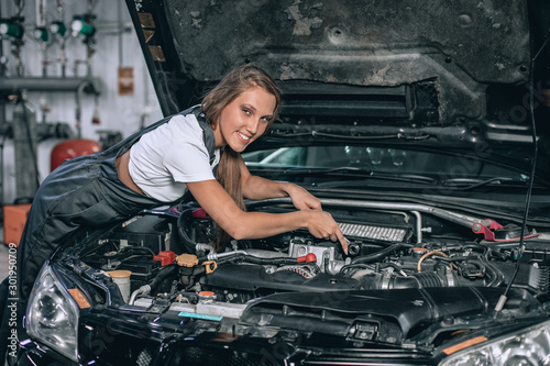 A brunette in a black jumpsuit and a white t-shirt near the open hood of black car. Young female in the garage is smiling at the camera. car repair concept