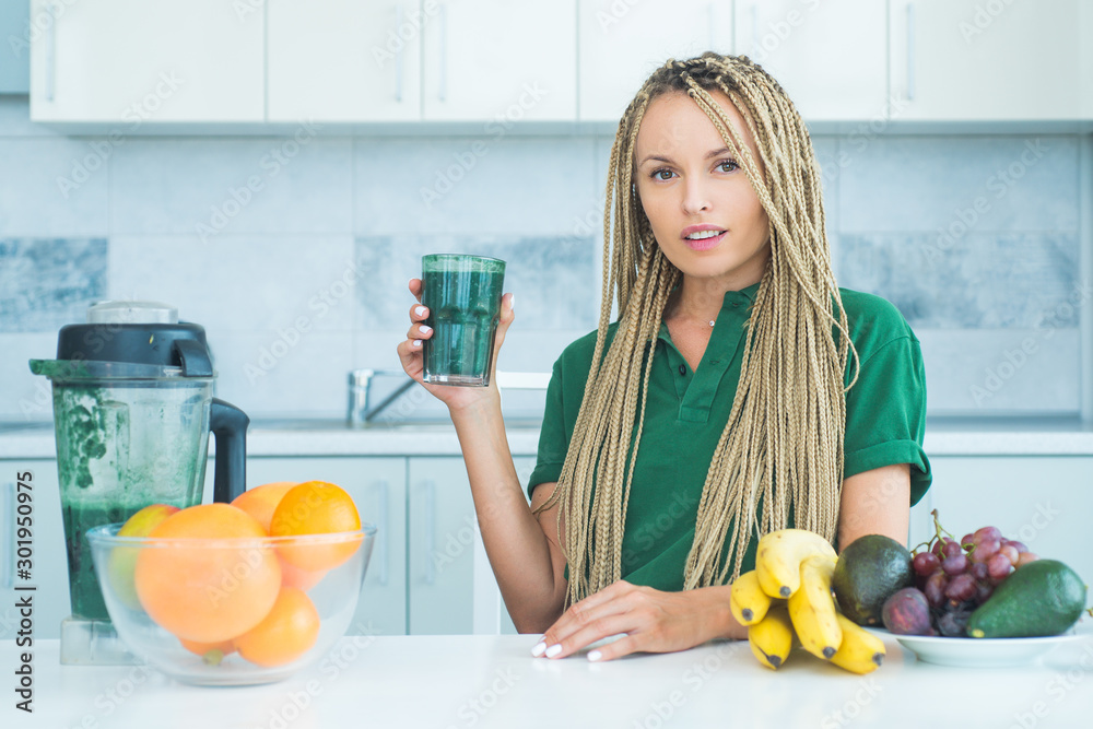 Positive woman hold cup of fresh green smoothie. Eco lifestyle. Young woman drinking green smoothie in the kitchen with fruits and vegetables. Vegan meal and detox menu. Positive woman hold cup of fresh green smoothie. Eco lifestyle. Young woman drinking green smoothie in the kitchen with fruits and vegetables. Vegan meal and detox menu.