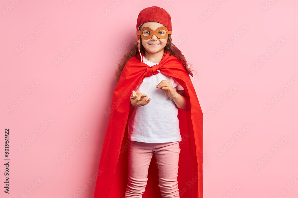 Portrait of caucasian kid girl wearing red cloak and helmet of pilot stand isolated over pink background. Girl hold wooden plane in hands