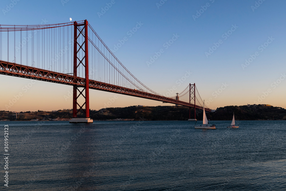 Sailing boats in the Tagus River passing by the 25 of April Bridge (Ponte 25 de Abril), in the city of Lisbon, Portugal; Concept for travel in Portugal
