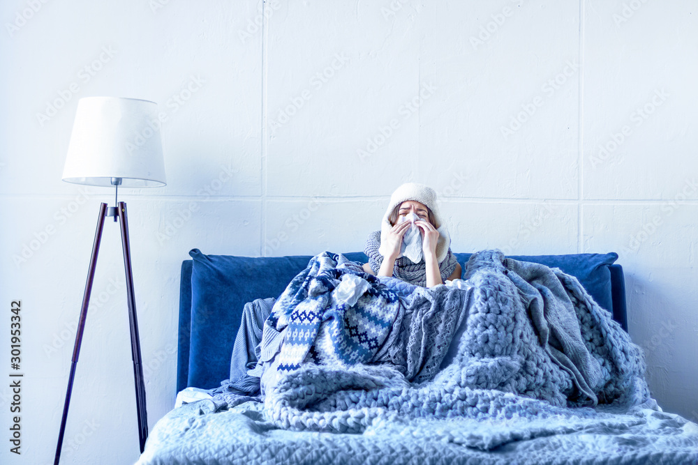 Sick exhausted girl in white warm hat wrapped herself in scarves and blankets is sitting in bed. Young woman with runny nose, fever and headache is treated at home. Winter cold and flu concept. - obrazy, fototapety, plakaty 