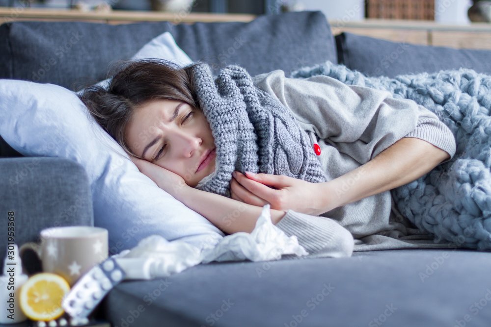 Sick exhausted girl in scarf is lying in bed wrapped in blanket. Young woman with fever and headache is measuring temperature with thermometer, treated at home. Winter cold and flu concept. - obrazy, fototapety, plakaty 