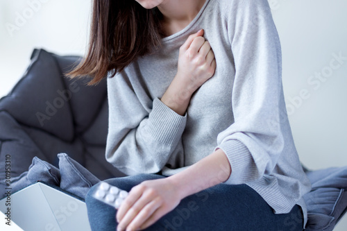 Young woman in grey clothes is holding hand on breast and going to take pills. Brunette girl is feeling bad. Sudden heart attack, myocardial infarction at home. Effect of stress concept. photo