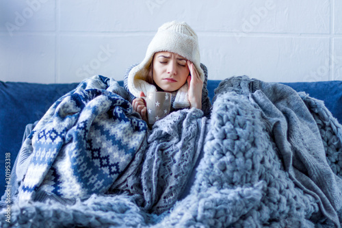 Tela Sick exhausted girl in white warm hat wrapped herself in scarves and blankets is sitting in bed