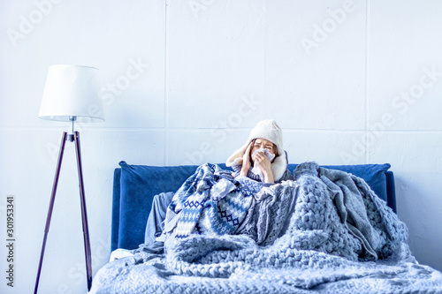 Fotografia Sick exhausted girl in white warm hat wrapped herself in scarves and blankets is sitting in bed