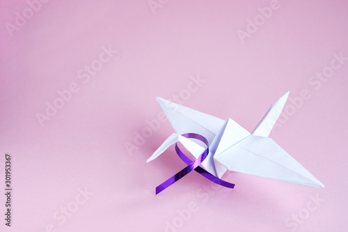 White paper crane with purple ribbon on pink paper background. Alzheimer's disease, Pancreatic cancer, Epilepsy awareness, hope, world cancer day. Hope concept. Copy space. Selective focus.  
