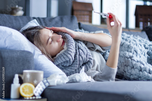 Sick exhausted girl in scarf is lying in bed wrapped in blanket. Young woman with fever and headache is measuring temperature with thermometer, treated at home. Winter cold and flu concept. photo