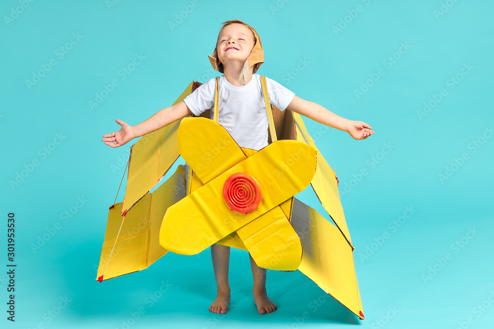 Dreams come true. Child boy represents how he flies at sky. Aviation future of kid wearing yellow toy plane, stand isolated oer blue background