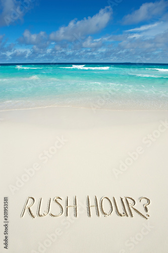 A humorous winter travel message 'RUSH HOUR?' handwritten on the smooth sand of a tropical island beach