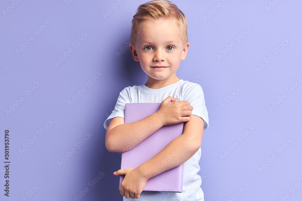 Little caucasian boy in white t-shirt stand holding purple bok isolated over purple background, looking at camera. Little caucasian boy in white t-shirt stand holding purple bok isolated over purple background, looking at camera.