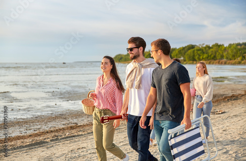 friendship, leisure and people concept - group of happy friends with guitar, picnic basket and folding chair walking along beach in summer
