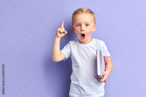 Caucasian child show bright emotions standing over purple background, holding purple notebook. With opened mouth
