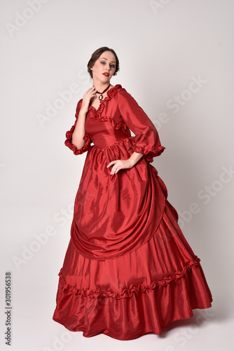full length portrait of a brunette girl wearing a red silk victorian gown. Standing pose on a white studio background.