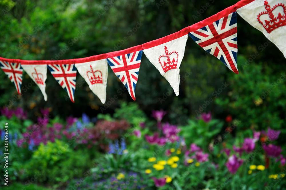 Banner of British Union Jack flag and royal crown celebratory bunting hanging in front of a flowery English summer garden background - obrazy, fototapety, plakaty 