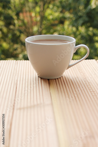 white cup with coffee on a light wooden table against a green summer garden, close-up, copy space