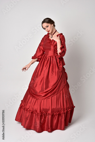 full length portrait of a brunette girl wearing a red silk victorian gown. Standing pose on a white studio background.