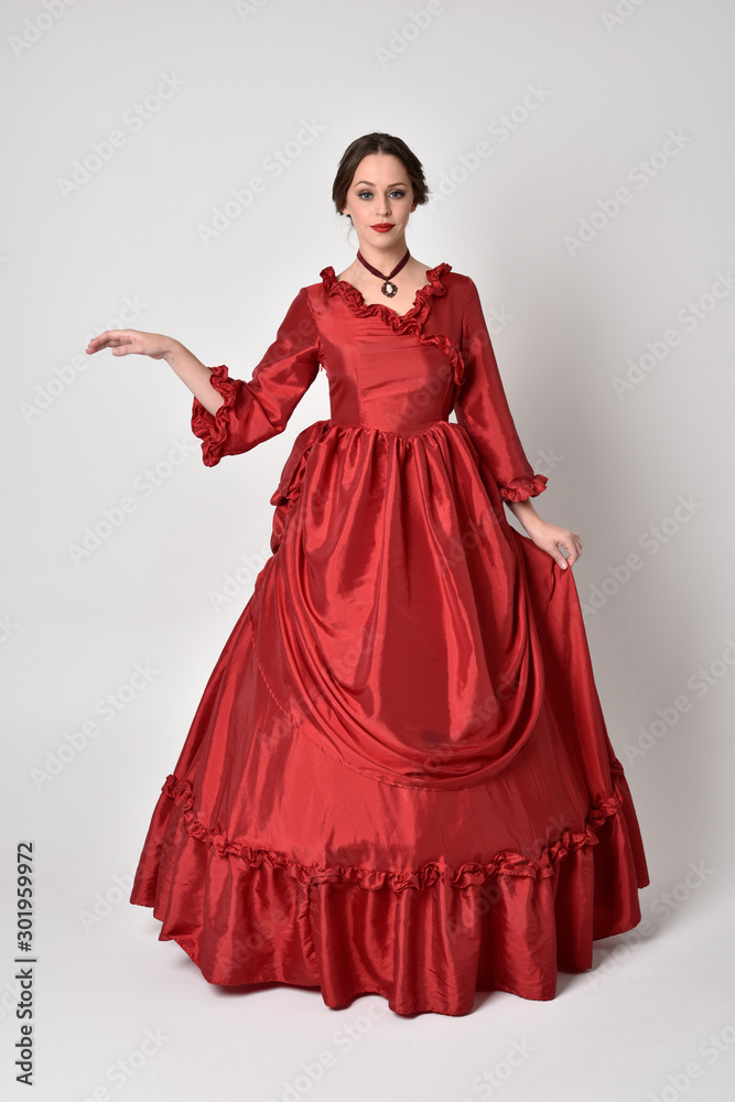 full length portrait of a brunette girl wearing a red silk victorian gown. Standing pose on a white studio background. full length portrait of a brunette girl wearing a red silk victorian gown. Standing pose on a white studio background.