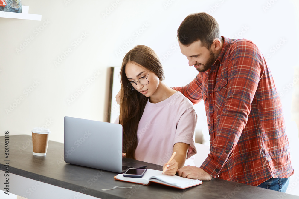 Attractive young female looking for important information in internet, unshaved man stands behind back with kind smile, background of white wall Attractive young female looking for important information in internet, unshaved man stands behind back with kind smile, background of white wall