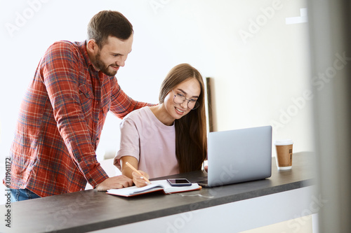 Pleasant happy girl with long hairs, in optical glasses looking at laptop computer with engaging smile, expresses positive emotion, happy to get good news