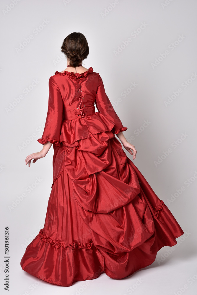 full length portrait of a brunette girl wearing a red silk victorian gown. Standing pose, with back to the camera, on a white studio background. full length portrait of a brunette girl wearing a red silk victorian gown. Standing pose, with back to the camera, on a white studio background.