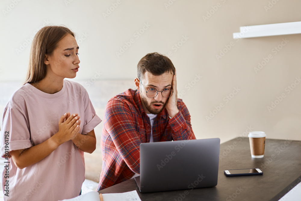 Worried young woman stands in brightly lighted room next to frustrated man, holds hands together on chest, prays for good game ending, hopes for miracle Worried young woman stands in brightly lighted room next to frustrated man, holds hands together on chest, prays for good game ending, hopes for miracle
