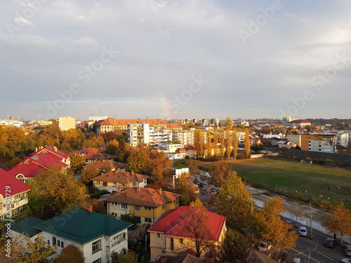a quarter of a rainbow over a city in Romania on a cloudy autumn day. many houses to the left of the frame and an old stadium to the right. Background wallpaper image