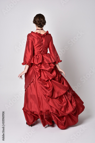 full length portrait of a brunette girl wearing a red silk victorian gown. Standing pose, with back to the camera, on a white studio background.