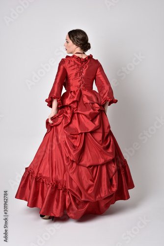 full length portrait of a brunette girl wearing a red silk victorian gown. Standing pose, with back to the camera, on a white studio background.