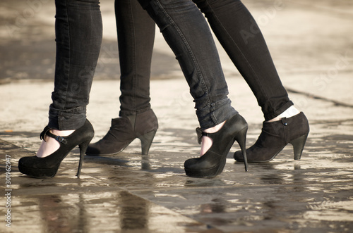 Unrecognizable young women walking in black skinny jeans and high heels across puddles on a sidewalk
