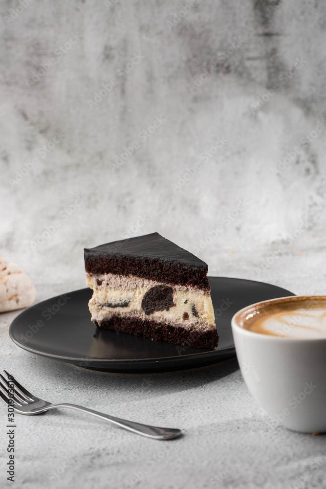 Creamy cheesecake with chocolate cookies and cream biscuits, chocolate cookies and cream biscuits cheese cake. Piece of cake on black plate, white cup on white marble background. Vertical photo.