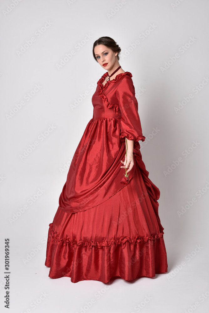 full length portrait of a brunette girl wearing a red silk victorian gown. holding a single rose on a white studio background.