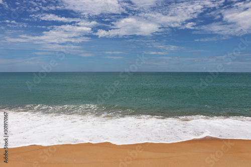 Surf on tropical sandy beach, scenic view to empty sea coast with yellow sand and emerald wave with white foam. Picturesque seascape with blue sky and white clouds