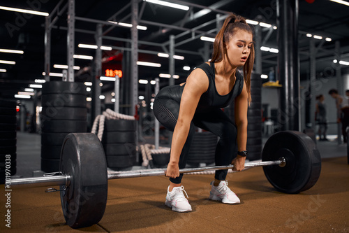 Young pretty fitness lady trying to lift heavy barbell from brown floor in modern gym hal, looking away with concentrated face, training hard for competition, indoor shot