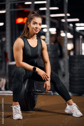Beautiful active woman with long straight hair, looking at camera with pleasant face, smiling cheerfully, having photo session in gym hall, sitting on barbell, sport bodybuilding powerlifting concept