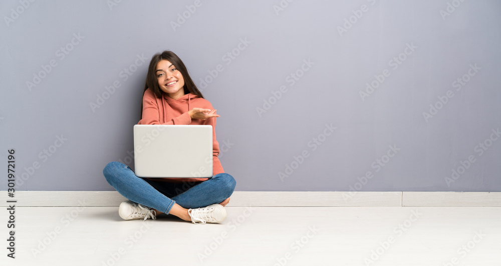 Young student girl with a laptop on the floor extending hands to the side for inviting to come - obrazy, fototapety, plakaty 