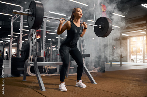 Charming pretty female athlete wearing black sportswear and white stylish sneakers, squatting in modern fitness studio using heavy barbell, white smoke in the air, portrait, selective shot