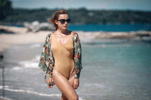 Young girl with a gorgeous body is resting on the beach with white sand near the ocean. Beautiful sexy model in a beige bathing suit and flower cape sunbathing.