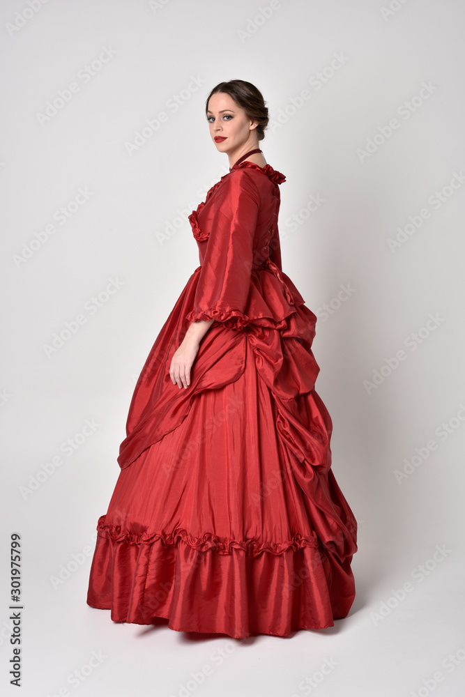 full length portrait of a brunette girl wearing a red silk victorian gown. Standing in side profile on a white studio background. - obrazy, fototapety, plakaty 