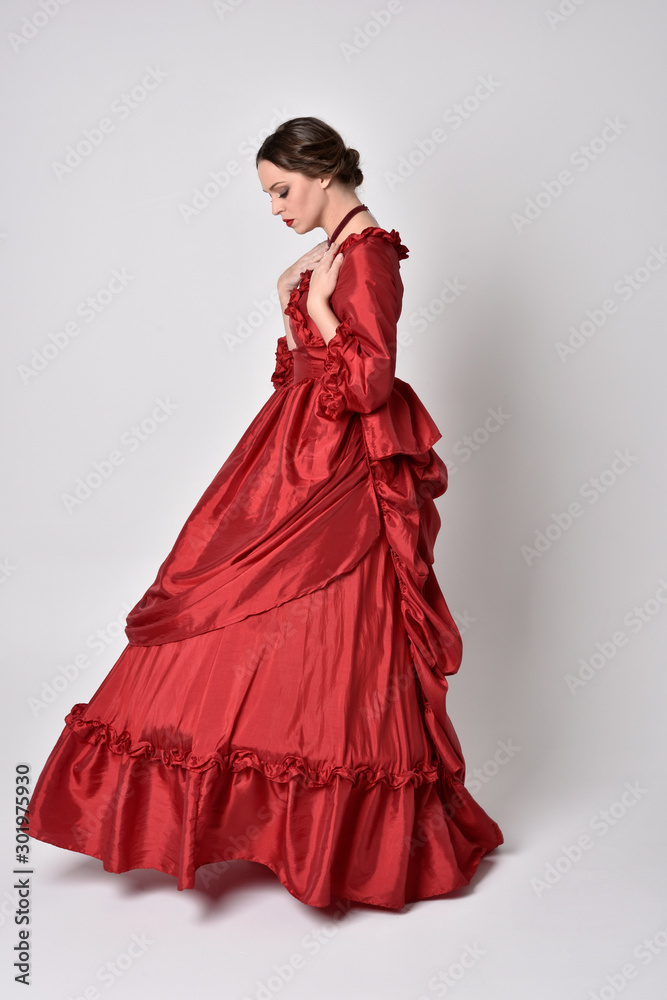 full length portrait of a brunette girl wearing a red silk victorian gown. Standing in side profile on a white studio background.
