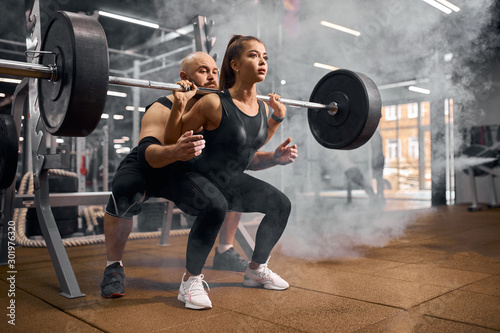 Pretty charming sports woman doing squats using heavy barbell, professional trainer standing behind, backing up, white smoke in the air, practicing in modern fitness center