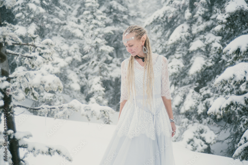Woman wearing elf ears, dreadlocks and white dress in winter snowy Christmas tree forest. Fog and mystery frozen day