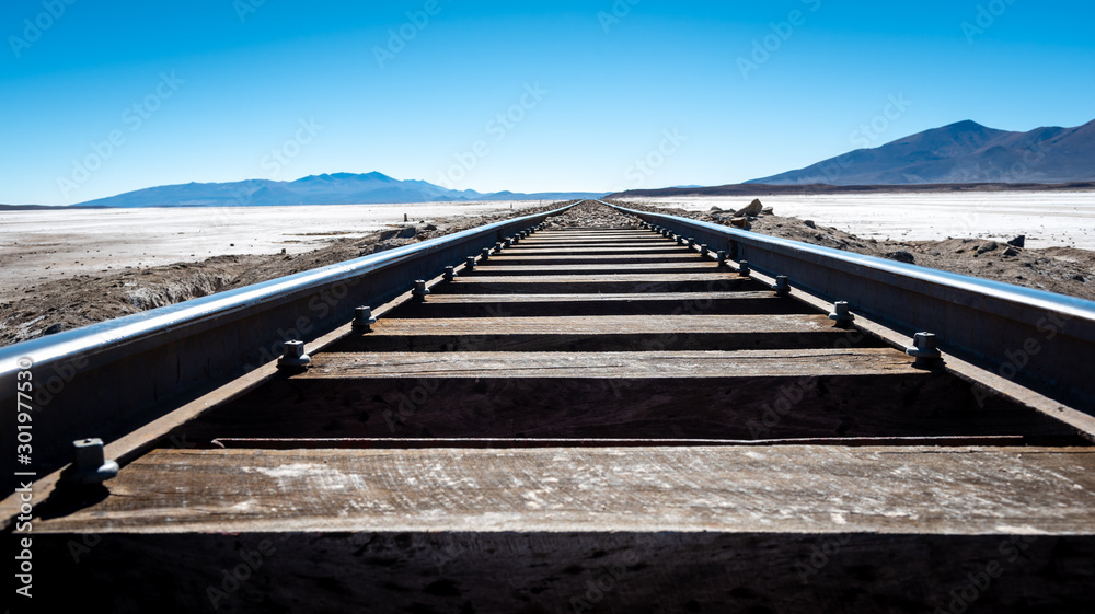 Wooden train tracks cross the Bolivian desert travelling towards the blue sky and distant mountains on the horizon.
