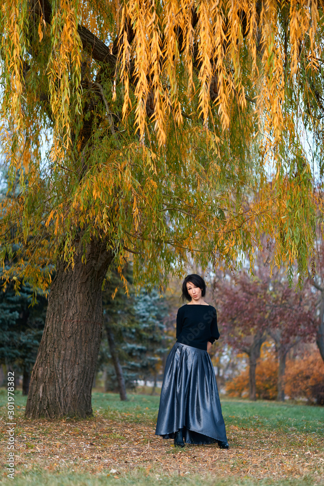 Beautiful elegant woman standing and posing near big yellow tree in a city park in autumn Beautiful elegant woman standing and posing near big yellow tree in a city park in autumn