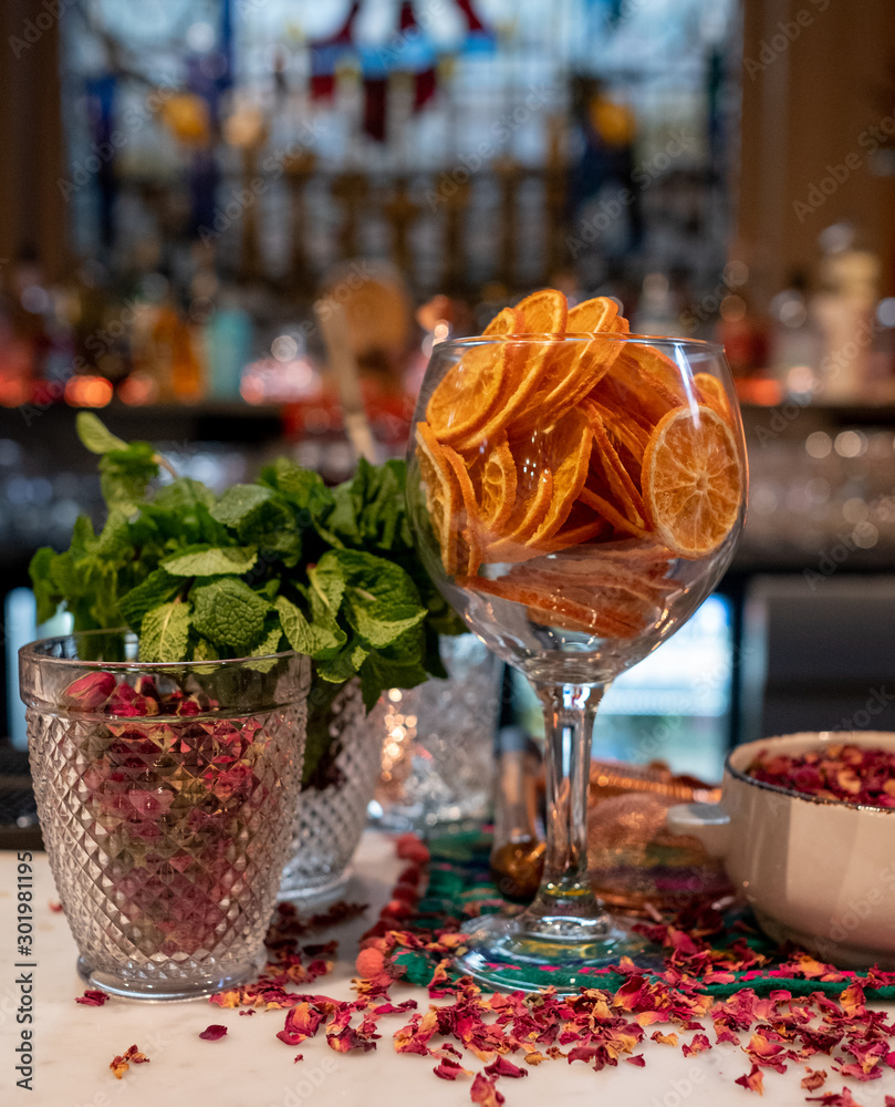 Glass full of dried sliced oranges, at a bar with rose petals sprinkled around for seasonal decoration. Bottles on shelves behind, out of focus.