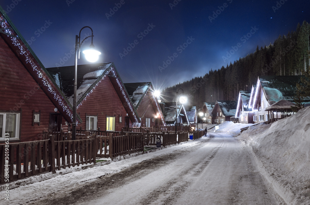 Fototapeta premium row of wooden houses on a snow-covered street in winter at night with dark sky